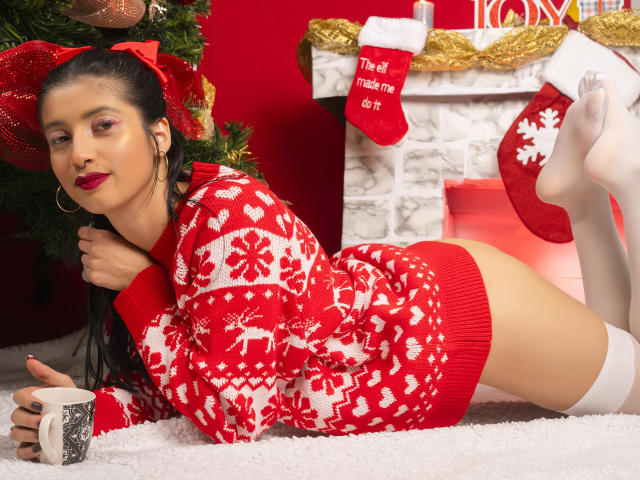 AlisKooper, with red sweater, white knee socks, and bare ass, lies on white carpet, holding a mug, beside a Christmas tree and fireplace. She has red lipstick and hoop earrings.