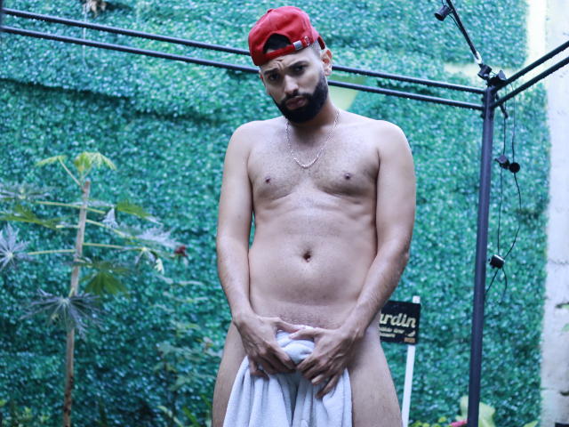 AntoniSmith desnudo, con un gorro rojo, sosteniendo una toalla blanca sobre su pene peludo, circuncidado y flácido. Tiene barba, piel de tono medio y un collar de cadena. El fondo es verde..