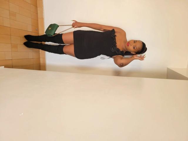 Freaky-hot woman in a black lace dress, holding a green chair, standing on wooden floor, wearing black knee-high boots, red lipstick, and dark hair in a ponytail.