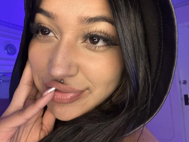 KatianPierce with dark hair, hooded, nose piercing, and lip piercing, smiling, touching her pink lips with white-manicured fingers, blue lighting, and long eyelashes.