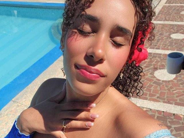Salma, con capelli castano ricci, un fiore rosso e rossetto rosa, occhi chiusi, mano al collo, indossa una maglietta blu a spalle scoperte, vicino a una piscina con piastrelle blu, alla luce del sole..