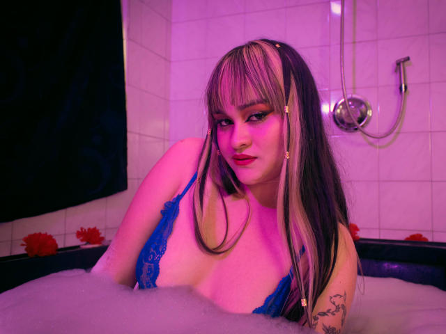 LailaDurmont in a bathtub with pink-purple lighting, wearing blue lace lingerie, showing her tits, with long black-and-blonde hair, and a tattoo on her right arm.