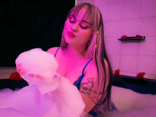 LailaDurmont with blonde hair, blue lace bra, holding bubble, pink lighting, tattoos on arm, sitting in bathtub, looking down with slightly parted pink lips.