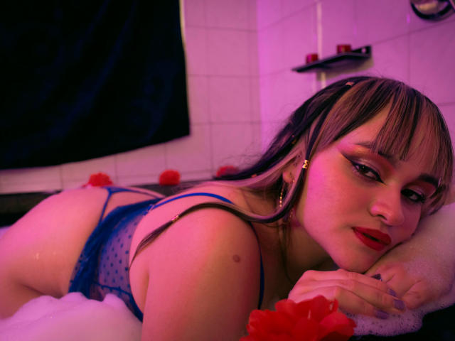 LailaDurmont lying in a bathtub, wearing blue polka dot lingerie, with red flower decorations, her ass up, long blonde hair with black streaks, red lipstick, and gold earrings.
