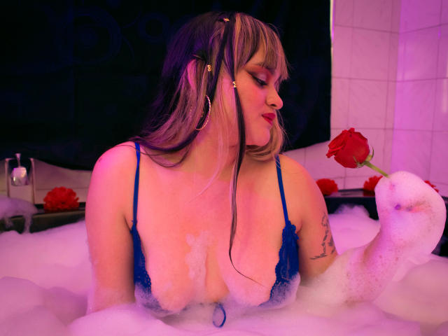 LailaDurmont in a bubble bath, wearing blue lace lingerie, holding a red rose, with her tits partially covered by foam, and her arm tattoo visible, under purple lighting.