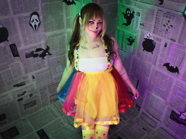 LailaDurmont with long brown hair, wearing a yellow, polka-dotted, strapless dress, white top, and yellow polka-dot stockings, has tattoos, green and purple lighting, and a playful smile.