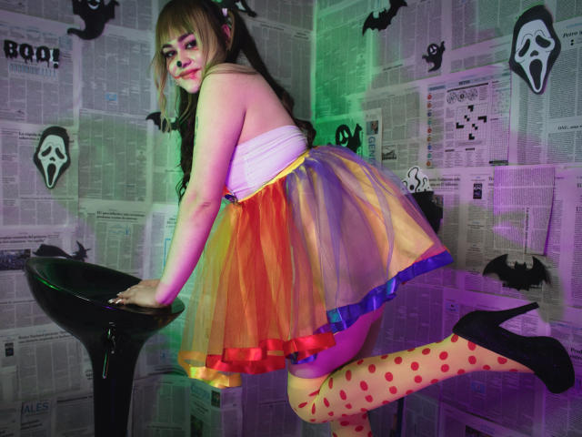 LailaDurmont with blonde pigtails, wearing a colorful sheer skirt, yellow polka-dot tights, black high heels, and a white strapless top, leaning on a black stool, showing her ass.