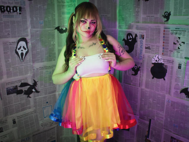 LailaDurmont with colorful rainbow skirt, white crop top, and suspenders, has puffy ass, small tits, tattoos, green and purple lights on her, long blonde hair with bangs.