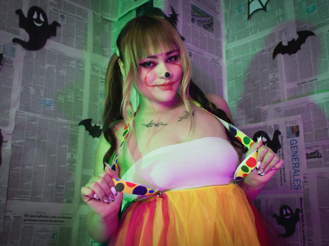 LailaDurmont with blonde pigtails, white strapless top, colorful polka-dot bra, yellow skirt, tattoos on chest, green and purple lighting, smiling, holding bra straps, in Halloween-themed setting.