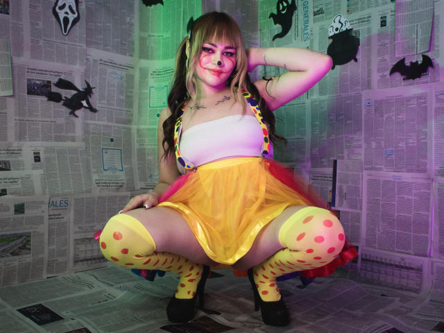 LailaDurmont crouching, wearing yellow polka-dot thigh-highs, yellow skirt, white top, and suspenders, with red panties, showing her ass, in a room with newspaper-covered walls.