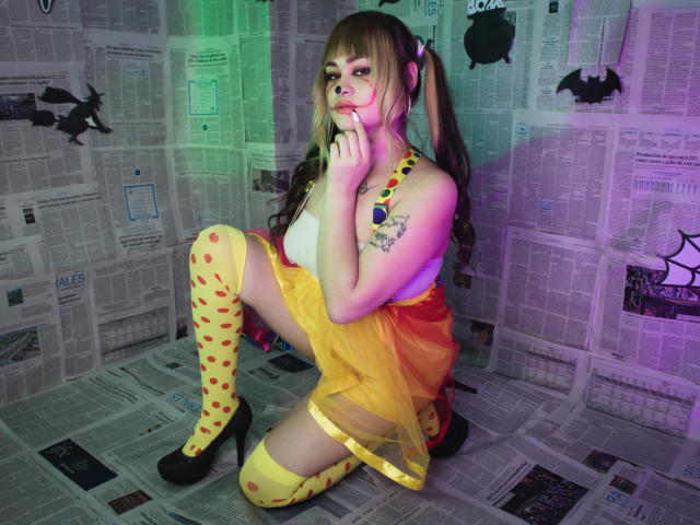 LailaDurmont, crouching, wearing yellow dress with red hearts, yellow thigh-highs, black heels, and pigtails, finger in mouth, showing tattoo on left arm, in colorful, paper-walled room.