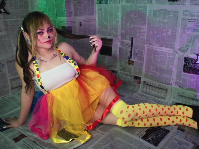 LailaDurmont with blonde pigtails, white tank top, colorful skirt, yellow polka dot thigh-highs, face paint, tattoos, leaning on newspaper-covered floor, holding hair, colorful skirt, big ass.