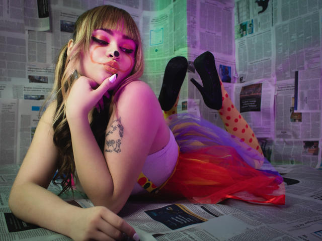 LailaDurmont with pink eyeshadow, pouty lips, and blonde pigtails, laying on newspaper, wearing a rainbow tutu, polka dot tights, and black heels, finger on her chin, tattoo on arm.