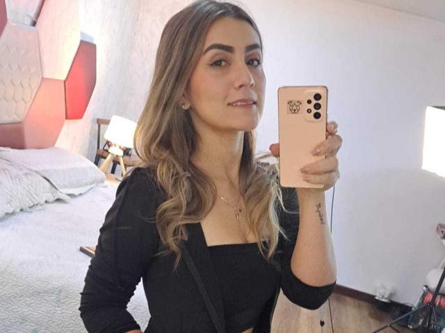 AriaRous with long, wavy brown hair, wearing a black top, holding a gold iPhone, taking a mirror selfie, showing a tattoo on her right forearm.
