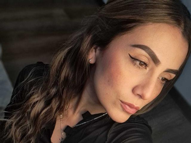 AriaRous with wavy brown hair, dark eyebrows, and winged eyeliner, wearing a black top, has a pensive expression, and a small pearl earring in her left ear.