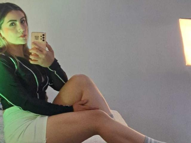 AriaRous with green and black long-sleeve top, white skirt, and white socks, taking a mirror selfie, showing her legs and butt, holding a gold phone.