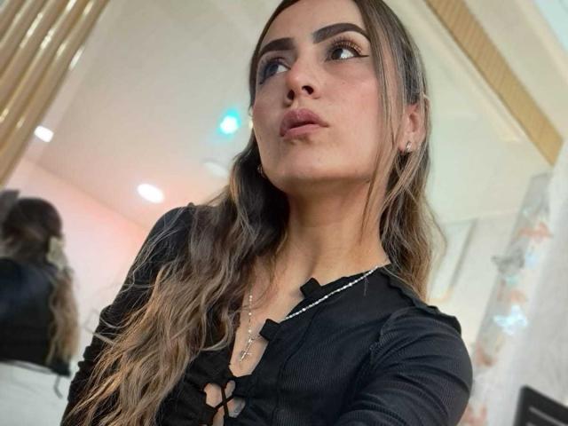 AriaRous with long brown hair, wearing a black top with cut-out design, pouty lips, looking up, silver necklace, mirror reflection in background, soft indoor lighting.