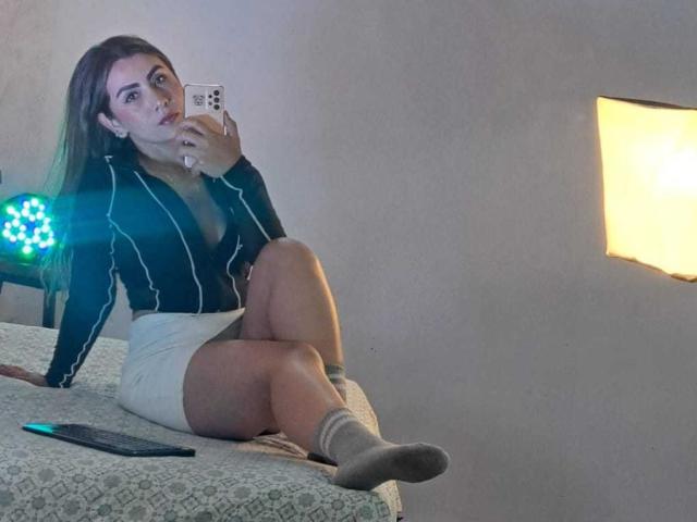 AriaRous with long brown hair, wearing a black striped shirt and white skirt, sitting on a bed, taking a mirror selfie, showing her tits and ass, in socks.