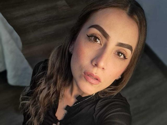 AriaRous has pouty pink lips, thick brown eyebrows, long wavy brown hair, and brown eyes. She's wearing a black shirt, looking up with a slightly open mouth.
