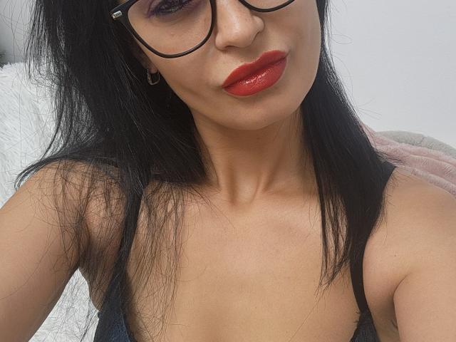 TheSunshine has long black hair, wearing black-framed glasses, and red lipstick. She has a medium skin tone and a slender build. Her top reveals a slight cleavage. The image is a close-up selfie, focusing on her upper chest and face. The texture of her hair is straight and slightly messy. The background is blurred and indistinct. The style is casual and contemporary. The sunlight highlights her features.