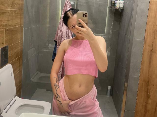 AmaiRin with dark hair, wearing a pink crop top, pulling down pink pajama pants, showing a tattoo on her right arm, taking a mirror selfie in a bathroom.