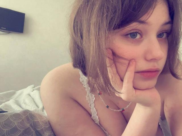 NikolKitt has light brown, shoulder-length hair, blue eyes, and is resting her chin on her hand, wearing a lace top that reveals her tits. She's looking at the camera.
