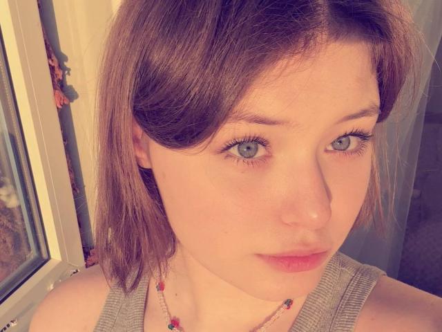 NikolKitt has light blue eyes, fair skin, and brown shoulder-length hair, wearing a gray tank top with a colorful necklace, looking directly at the camera with a slight smile.