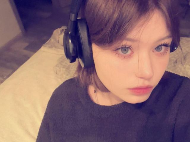 NikolKitt has light skin, blue eyes, and brown hair, wearing black headphones and a black sweater, looking slightly annoyed. Her small tits are partially visible.