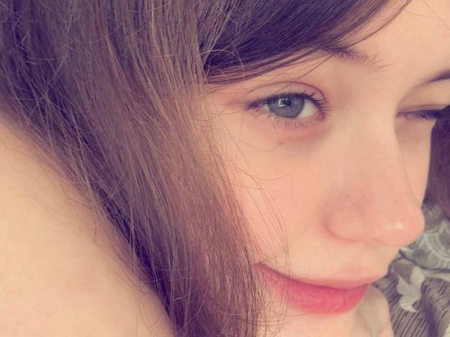 NikolKitt's face, showing blue eyes, pink lips, and a small mole near her right eye. Her brown hair is partially covering her left cheek. Her nose is slightly upturned.