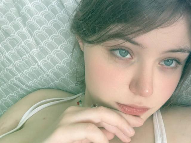 NikolKitt has pale skin, blue eyes, and brown hair, lying down with a pensive look, wearing a white tank top, her hand near her chin, lips slightly parted.