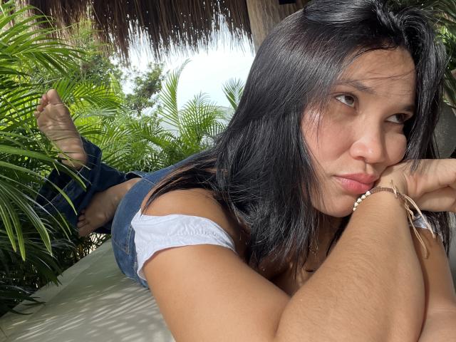 AnnySquiirt with black hair, lying on her stomach, wearing a white tank top and blue denim shorts, showing her bare ass, looking at the camera with pouty lips.