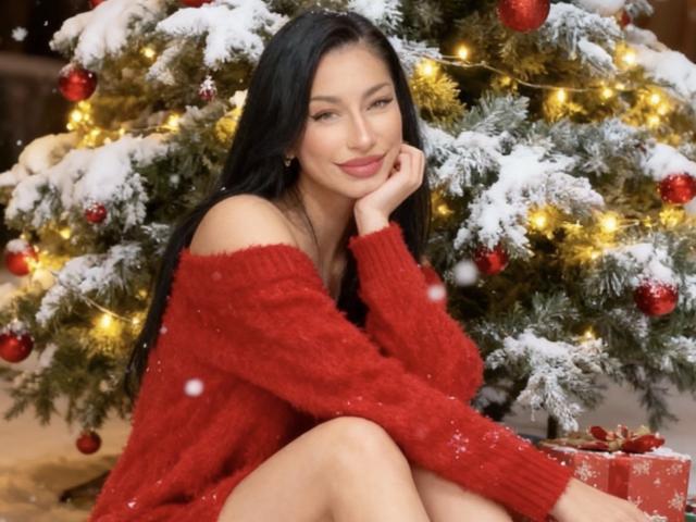 VenussXx with long black hair, red off-shoulder sweater, and cute smile, sitting by a snow-covered Christmas tree with red ornaments and yellow lights. Boobs slightly visible.