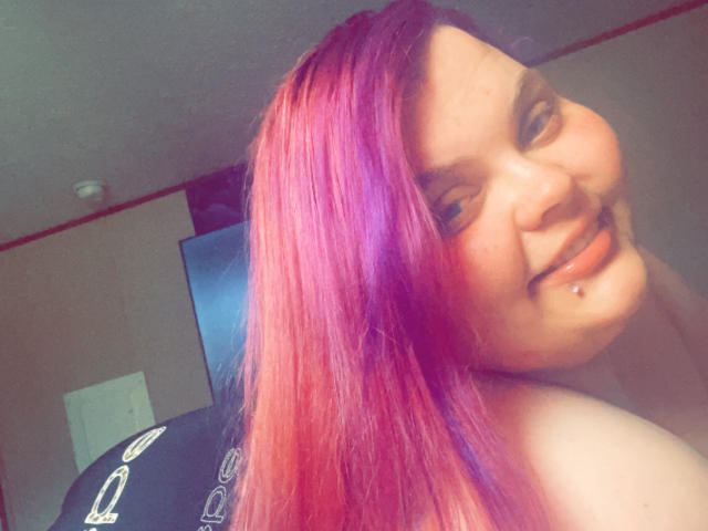SpoiledHazel with pink-purple hair, smiling at the camera, showing a lip piercing, looking over her shoulder, bare shoulders visible. Soft light, casual, relaxed.
