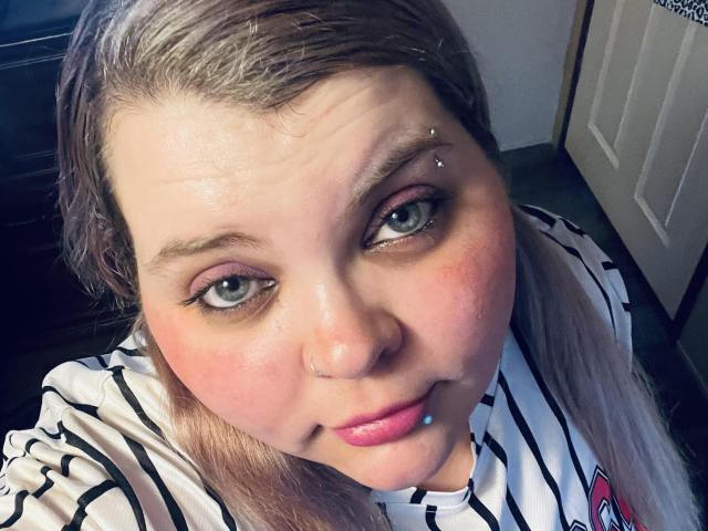 SpoiledHazel has light blue eyes, a nose piercing, and a lip piercing. She's wearing a striped shirt with pink lipstick, and her blonde hair is parted.