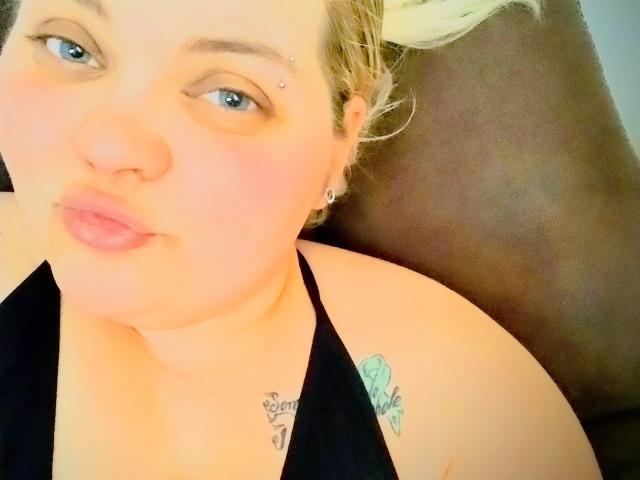 SpoiledHazel with light skin, blue eyes, and blonde hair, pouty lips, black halter top, small nose piercing, tattoo on right shoulder, lying on brown leather.