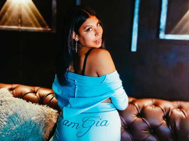 SashaLia with dark hair, looking over her shoulder, wearing a blue off-shoulder dress, exposing her black bra strap, and a fluffy pillow on a brown leather couch.