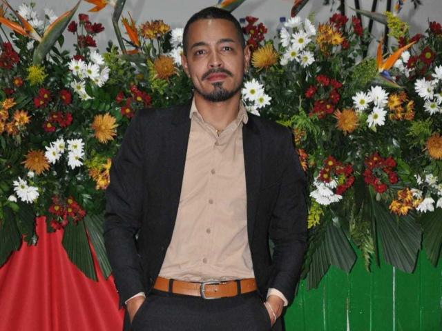 AlexCarls stands with hands in black pants pockets, wearing a tan shirt and black jacket, in front of a colorful floral arrangement with white, yellow, and red flowers.