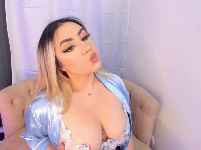 MelissaLust with blonde hair, pouty lips, and heavy eyeliner, wearing a blue satin robe, exposing her big tits with lace, sitting on a pink chair.