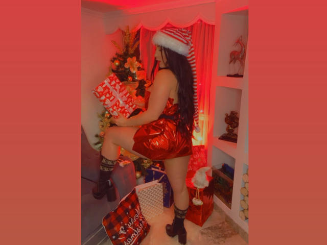 AgathaRuiz with long black hair, red Santa hat, shiny red dress, black boots, holding presents, standing on one leg, showing her ass, in a red-lit room.