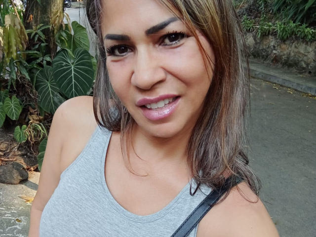 BrigiteWalsh has medium brown skin, straight brown hair, and is wearing a grey tank top showing her small tits. She's smiling with pink lipstick and long eyelashes.