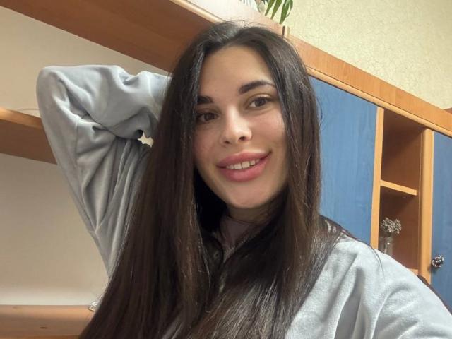 BlackFatal, con lunghi capelli castani, felpa grigia, braccio alzato, sorridente, con labbra rosa e occhi marroni. Lo sfondo presenta scaffali in legno e armadietti blu..
