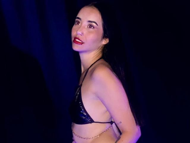 MeganJhonsom with pale skin, red lipstick, and long black hair, wearing a shiny black bikini top, chain belt, and smiling at the camera, showing her tits.
