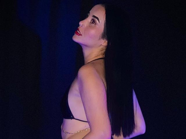 MeganJhonsom with long black hair, red lipstick, black bra, chain belt, and exposed back, looking over shoulder, in dim blue-purple light. Boobs visible.