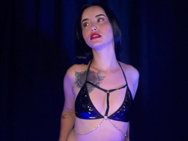 MeganJhonsom with black shiny bra, chain belt, and tattoos, looking up with red lips, short black hair, and a slight smile. Boobs are visible, tits are small to medium.