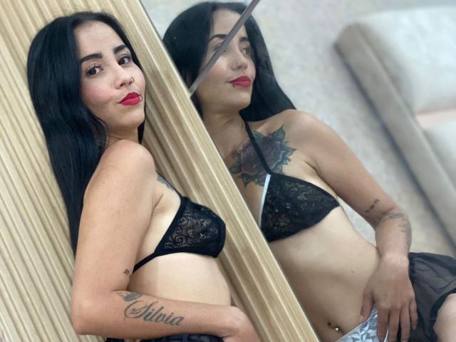 MeganJhonsom in black lace bra, showing her tits, with a reflection, has long black hair, red lipstick, tattoos, and a belly button piercing, standing against a mirror.