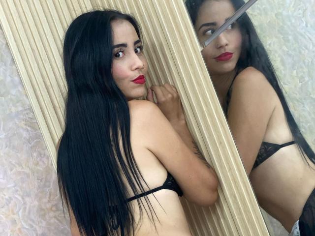 MeganJhonsom with long black hair, red lipstick, and black lace bra, standing sideways in front of a mirror, showing her back and reflection of her tits.