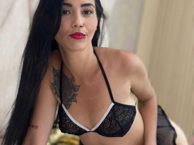 MeganJhonsom with long black hair, red lipstick, black lace bra, small tits, tattoo on left chest, nose ring, leaning forward, smiling, bedroom setting.