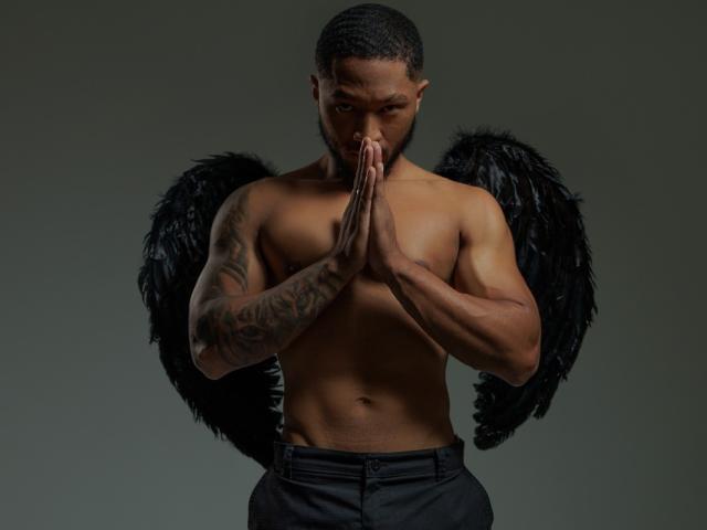 JadenSmithy's muscular, tattooed chest with black angel wings, standing shirtless, hands in prayer position, serious expression, dark skin, short beard, black pants, gray background.