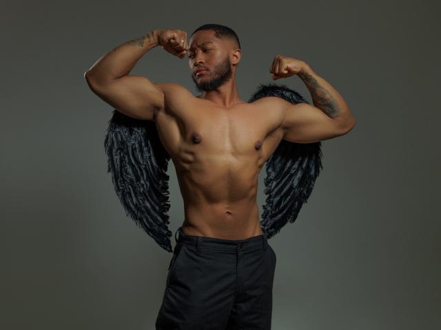 JadenSmithy, muscular, shirtless, flexing biceps, black angel wings, black pants, tattoo on right arm, short beard, serious expression, dark skin, studio lighting, gray background.