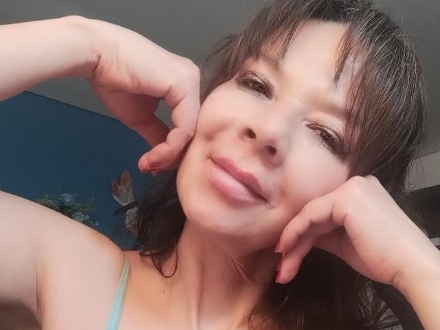 SamaraBellet with hands on cheeks, smiling slightly, brown hair, blue tank top, fair skin, subtle makeup, brown eyes, soft light, casual selfie, hands up, gentle expression.
